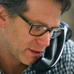 A man with glasses talks on the phone, wearing a blue and white checkered shirt.