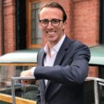 A person in a suit and glasses stands outdoors, leaning on a car door, with a brick building in the background.