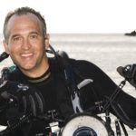A person in scuba gear holding an underwater camera, with the ocean and a rocky outcrop in the background.