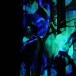 Three people stand in a dimly lit room with blue and green light projections creating abstract patterns on them and the wall.