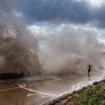 Huge waves crashing onto a coastal road as a person runs to escape the spray, under a cloudy sky.