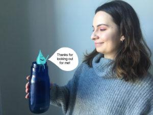 A woman in a gray sweater holds a dark blue water bottle, smiling at a water droplet character sitting on its lip. The droplet says "Thanks for looking out for me!"