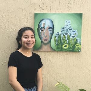 Katharine Guzman smiling as she stands by her painting hanging on a wall.