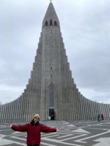 Ely standing in front of the point-shaped Hallgrímskirkja church in Reykjavík with arms outstretched.