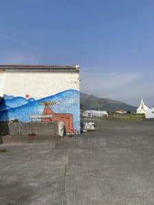 Ely's second DANDA mural, featuring the character sitting naked in front of blue mountains, a red sun sitting in a white sky. The largest mountain in the mural connects with the real mountain of the landscape behind the building.