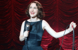 Rachel Brosnahan as Mrs. Maisel, smiling as she holds a mic in one hand and a mic stand in the other.