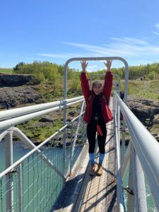 Ely standing on a metal bridge crossing a river, her arms stretched out above her to touch a metal pole running across the top.