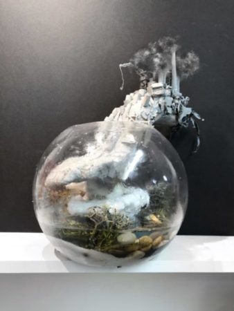 3D artwork. Clear crystal ball filled with water, rock, paper, and trash. Coming out of the top of the sphere is a cityscape with towers billowing pollution into the air.