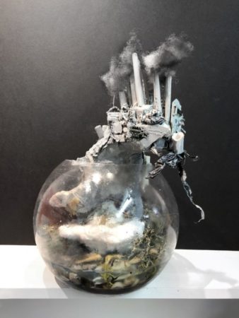 3D artwork. Clear crystal ball filled with water, rock, paper, and trash. Coming out of the top of the sphere is a cityscape with towers billowing pollution into the air.