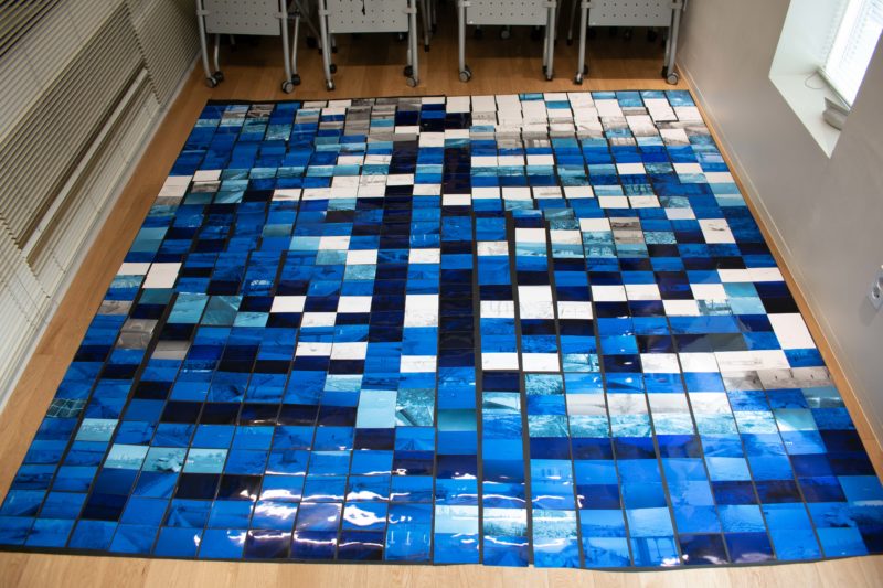 Installation comprised of many photographs of a river. The photos are varying shades of blue or white. The effect is as if the floor were made of river photos.