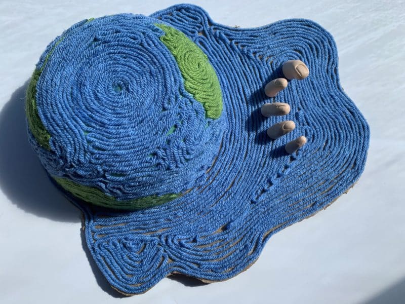 3D. An ocean made of yarn. Within the ocean is a half globe (Earth). Beside it are the figures of a hand coming out of the ocean surface.
