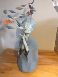 A clay sculpture of a seated figure with flowing hair, decorated with colorful leaves. The figure wears a calm expression and has intricate patterns on its face.