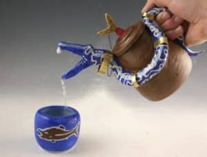 A hand pours water from a brown teapot with a blue, fish-shaped spout into a blue cup decorated with a fish design.