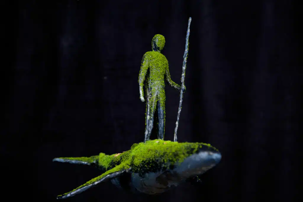 A moss covered man stands on a whale holding a staff.