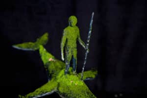 A small figure covered in green moss stands holding a stick on the back of a moss-covered fish against a dark background.