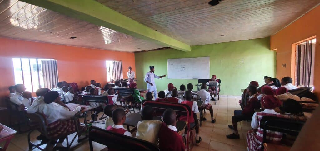 A teacher stands at a whiteboard, instructing a classroom filled with students in uniforms, some with red caps, in a room with orange and green walls.