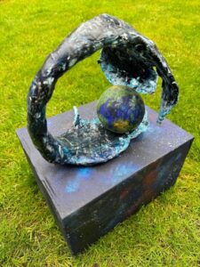 Abstract sculpture with a circular frame encases a spherical object on a black rectangular base, set on grass.