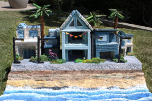 A miniature model of coastal houses with palm trees, colorful flags, and a pink bicycle on the street. The setting includes a painted ocean and sandy beach in the foreground.
