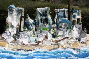 A miniature model of a coastal town with white buildings, some in ruins, set against a backdrop of waves and greenery.