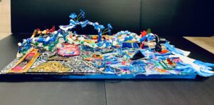 Colorful abstract sculpture with winding blue structures, floral elements, and various textures on a flat surface, set against a plain dark background.