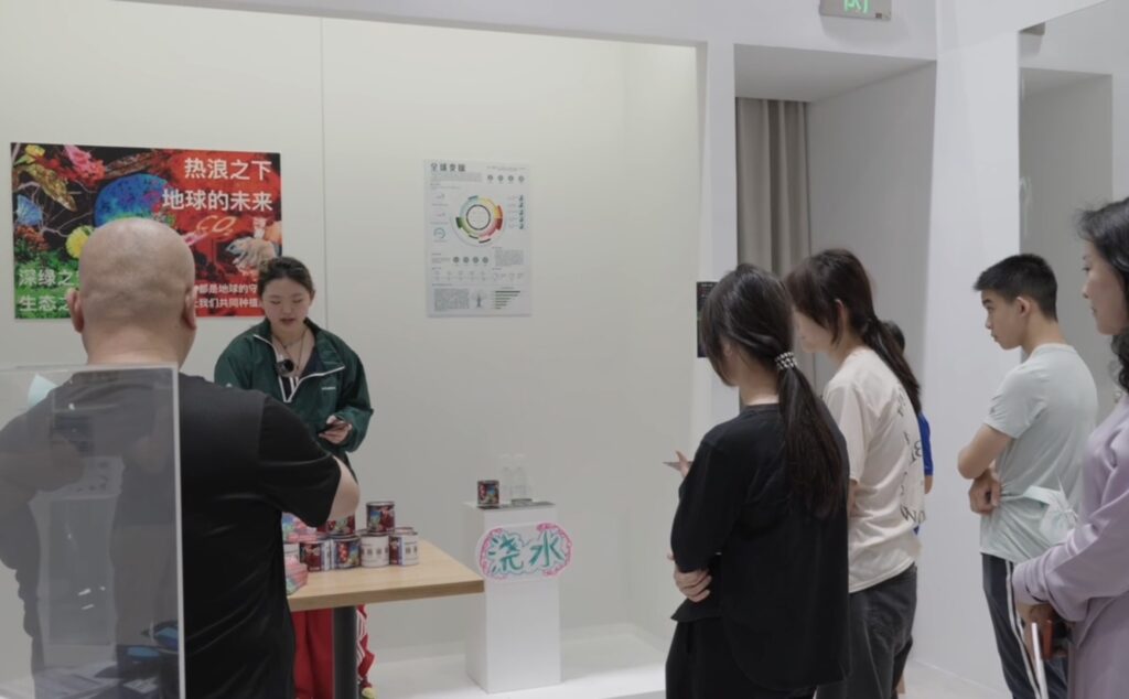 A woman stands behind a table giving a presentation to several people in a room with informational posters on the walls.