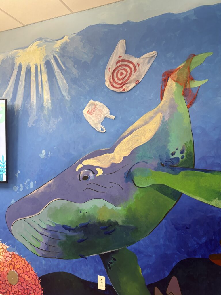 A mural of a whale underwater with plastic bags floating above it. The sun shines through the water.