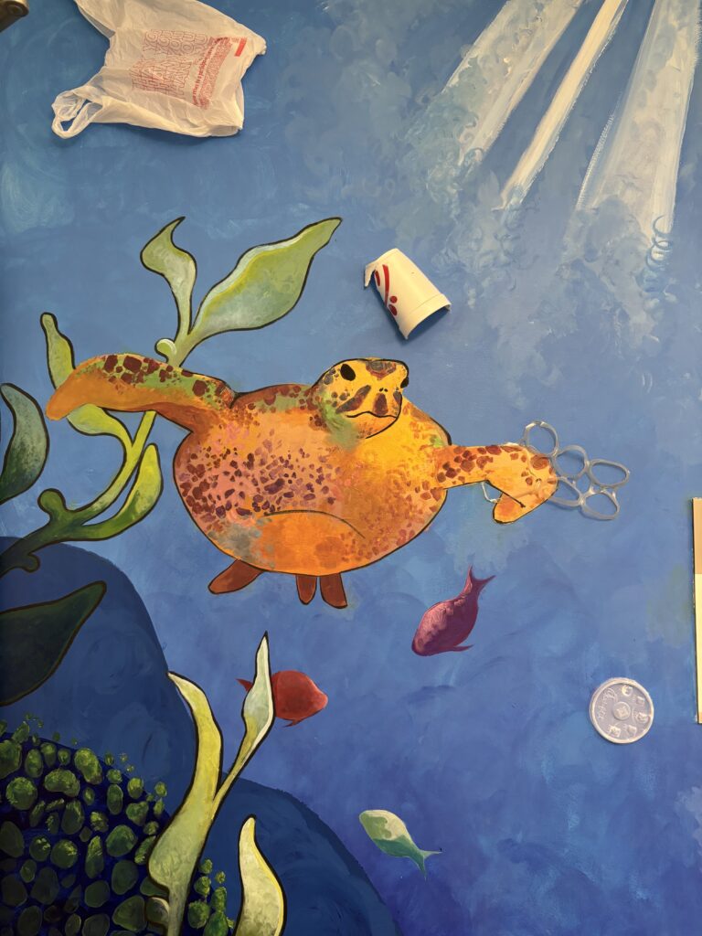 A colorful mural of a sea turtle swimming underwater, surrounded by fish, with plastic debris like a bag, cup, and rings above it. Sunlight filters through the water.