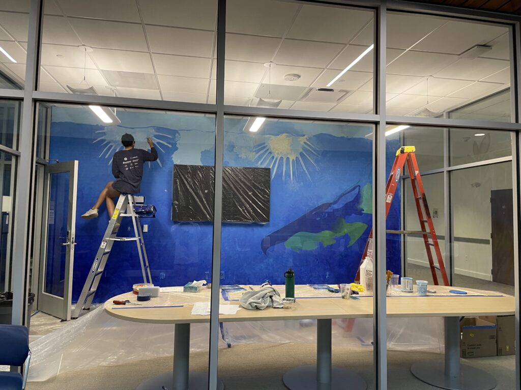 Person on a ladder painting a mural on a blue wall in a room with large windows. Paint supplies and plastic coverings are on the table and floor.